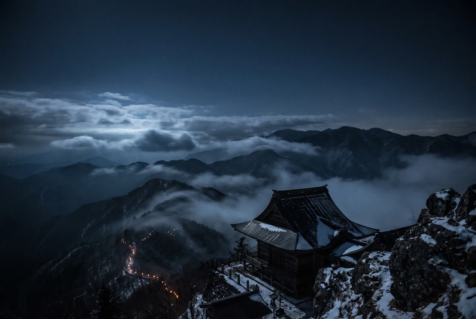 A summit clearing above the temple, looking back down across ridgelines to distant amber lantern lights.